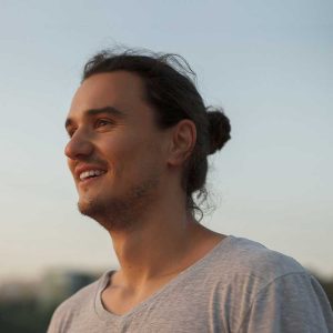 Profil młodego mężczyzny z brodą i włosami spiętymi w kok, ubranego w szary t-shirt. Mężczyzna uśmiecha się szeroko, patrząc w dal na tle jasnego, rozmytego nieba o zmierzchu.