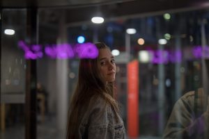 Młoda kobieta o długich włosach patrzy przez ramię w stronę aparatu. Znajduje się w nowoczesnym, przeszklonym budynku wieczorem, co tworzy nastrojową atmosferę z odbiciami fioletowych neonów na szybie.
