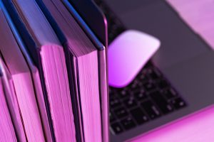 Zbliżenie na dłonie osoby w beżowej sukience, która trzyma otwarty notes i długopis, przygotowując się do robienia notatek. W tle widać niewyraźną postać innej osoby siedzącej na kanapie, co sugeruje sesję terapeutyczną lub wywiad.