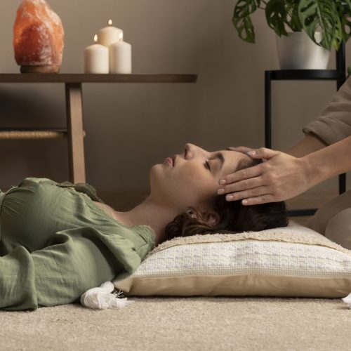 Kobieta leży na macie z zamkniętymi oczami podczas sesji relaksacyjnej. Terapeuta siedzący obok kładzie dłonie na jej głowie. W tle na półce znajdują się palące się białe świece oraz lampa solna, co podkreśla atmosferę spa.